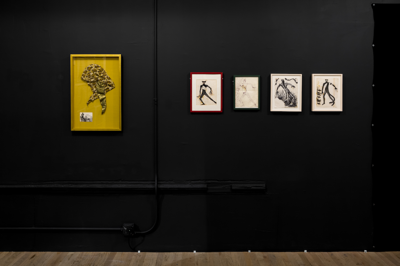 Several framed artworks hanging on a black gallery wall. A large yellow frame hangs on the left side of the wall while on the right, four smaller anthropomorphic drawings are displayed.