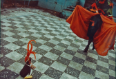 A film photograph of a person in a black dress and red cape, advancing across a checkered floor towards a small plastic penguin wearing beaded jewelry and a red feathery crown.