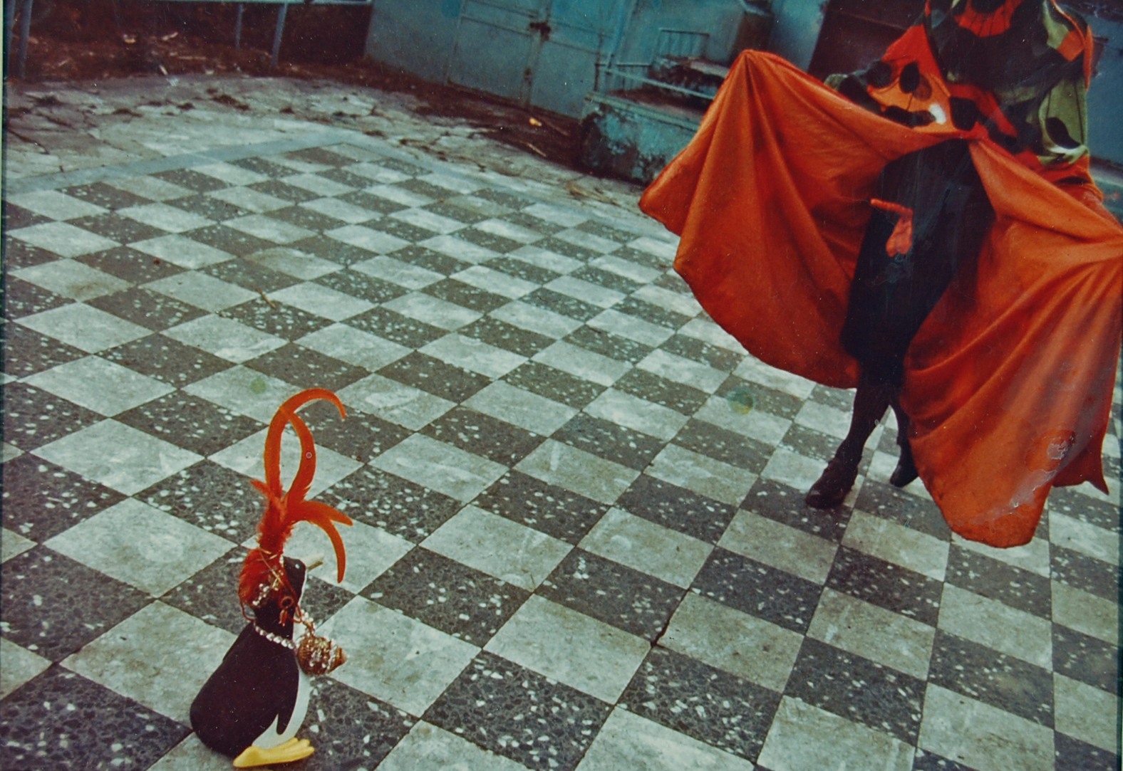 A film photograph of a person in a black dress and red cape, advancing across a checkered floor towards a small plastic penguin wearing beaded jewelry and a red feathery crown.
