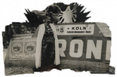 A black and white image with an organic border features Jack Smith in a white cloak standing in front of a wall. To the left of him are two posters that read, 