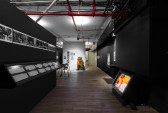 Black and white photographs hang in a horizontal line across a dark wall descending into an exhibition space. On the right, a video monitor sits on the ground. In the background is a free-standing wall featuring a series of framed photographs. A mannequin dressed in orange and green fabric is displayed in front of the photographs.