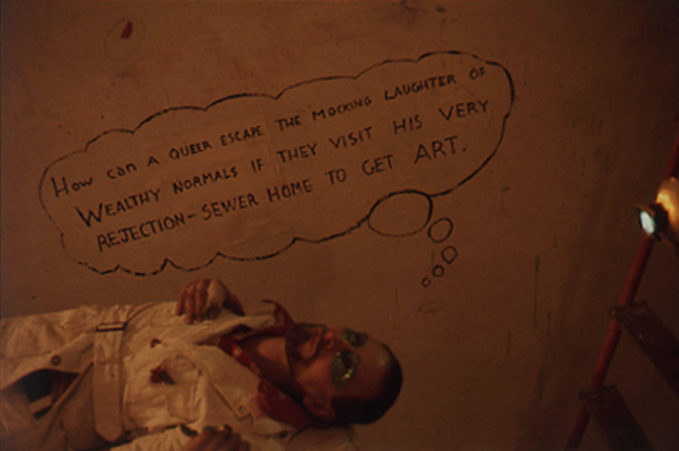 A person, clad in a white jumpsuit and blue eye makeup, lies horizontally on the ground. Their top half body is visible in the bottom left corner. Next to them floats a handwritten thought bubble, which reads, 
