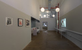 A gallery space with high ceilings and wooden flooring. On the left, three paper collages hang on a two-toned wall. Two videos are projected onto the right wall side-by-side. In the background, a doorway frames a black and white, projected image of a man's face.