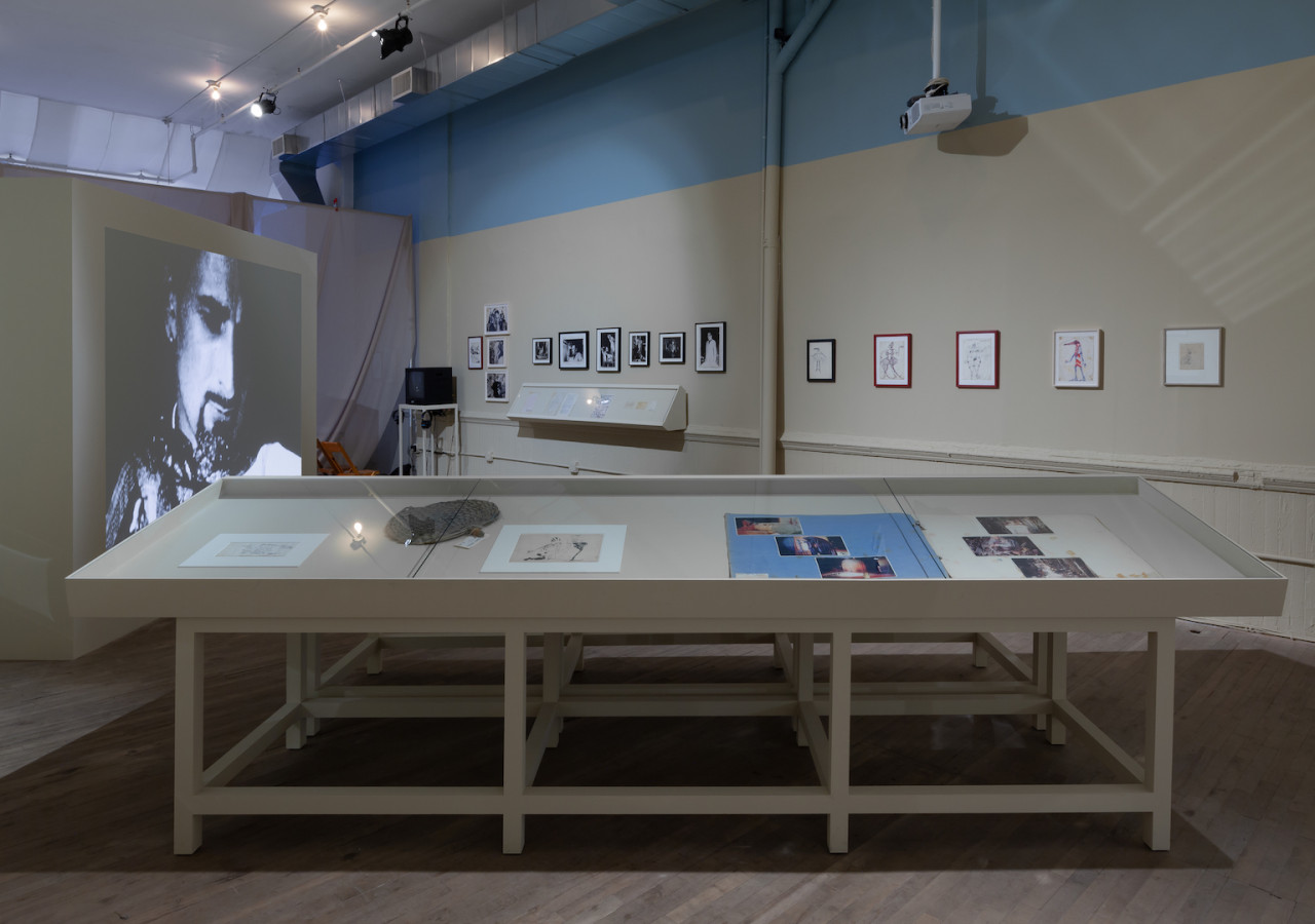 A display case holds materials in the middle of a neutral colored room. A black and white video is projected on a wall on the left side of the room. The projection faces a wall displaying several framed photographs and drawings.