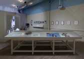 A display case holds materials in the middle of a neutral colored room. A black and white video is projected on a wall on the left side of the room. The projection faces a wall displaying several framed photographs and drawings.