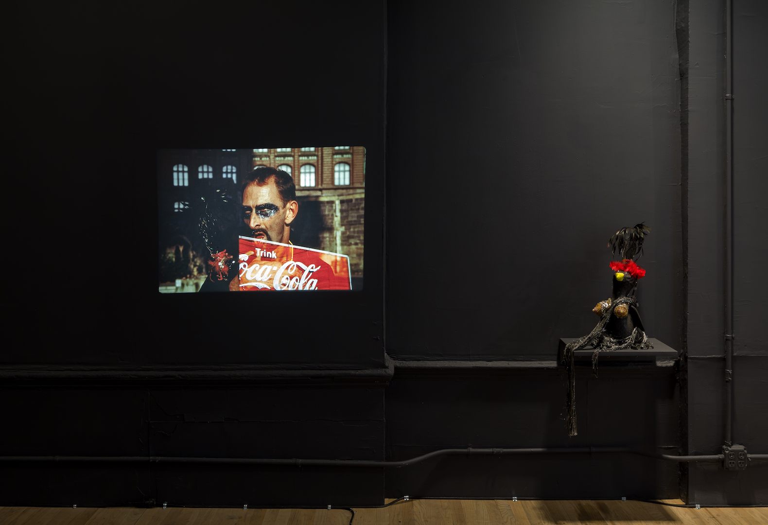 An image of a man with black material painted around his eyes projected onto a black gallery wall. A portion of a red Coca-Cola sign reading 