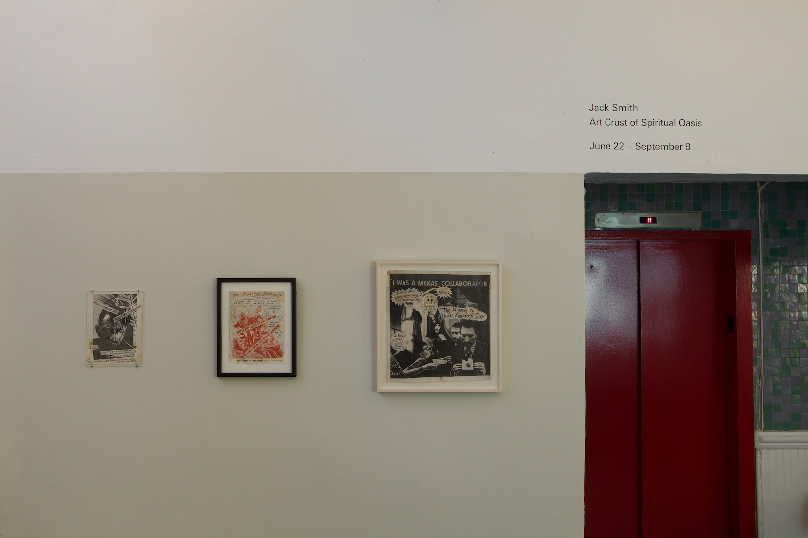 A two-toned wall with three paper collages hanging next to a niche that houses a red elevator. Vinyl text displayed on the wall above the niche reads, 