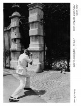 A black and white image of a man photographing a plastic penguin in front of a decorative gate. Sideways text on the right reads, 