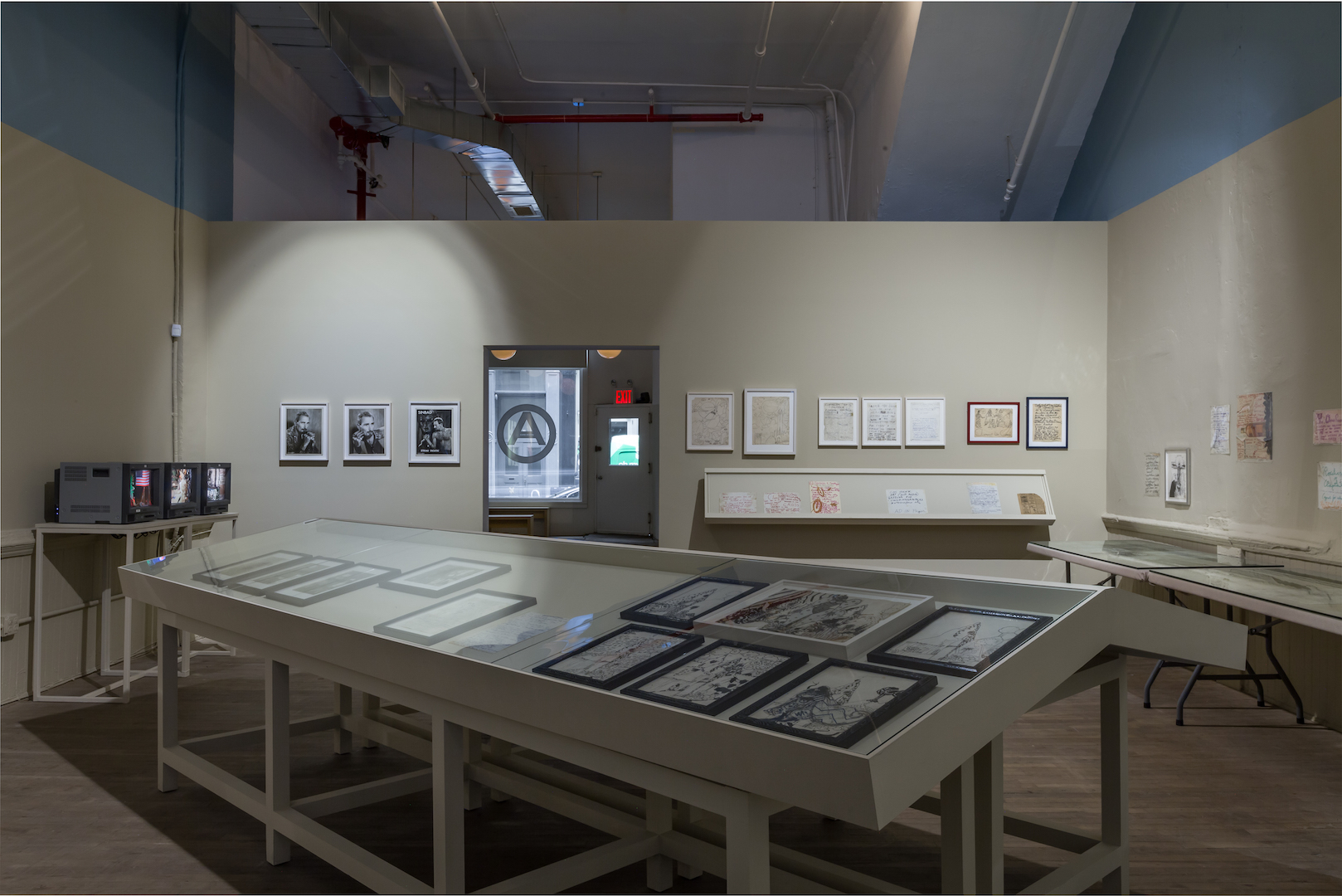 An angled view of a display case containing several framed drawings. To the left of the display case, three vintage television sets rest a white support and play video footage. Framed documents and photographs hang along the back wall on either side of a doorway.