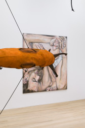 A partial view of a large, orange circular slug sculpture is in front of a painting hanging in a white-walled gallery space.