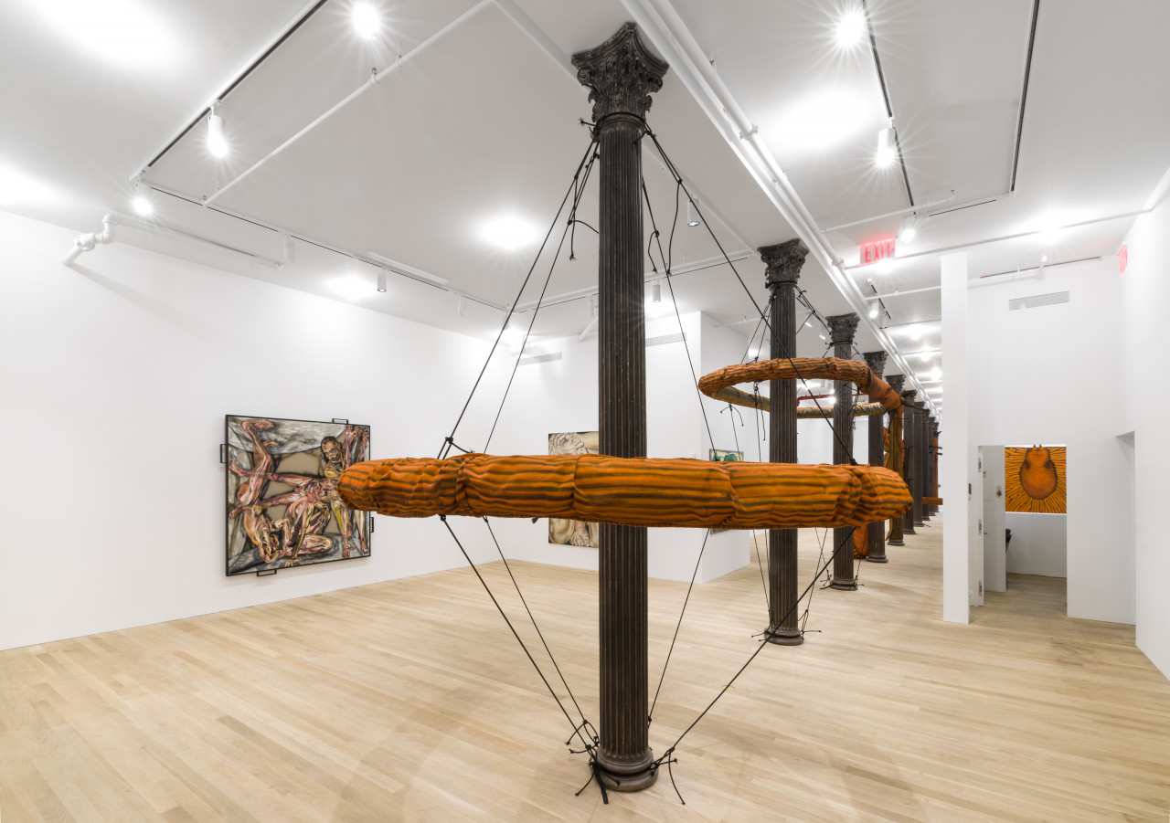Two large, orange slug sculptures forming circular ring shapes around cast-iron columns suspended by elastic cords are visible inside a white-walled gallery space along with square paintings on walls in the background.