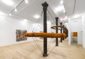 Two large, orange slug sculptures forming circular ring shapes around cast-iron columns suspended by elastic cords are visible inside a white-walled gallery space along with square paintings on walls in the background.