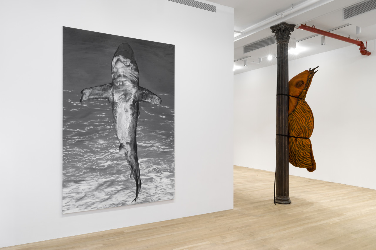 In the left foreground of the image is a painting of a shark standing upright in water and in the right background of the image is a large, orange stuffed slug sculpture strapped to a cast-iron column in a white gallery space.