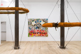Two large, orange slug sculptures forming a circular ring shape around separate cast iron columns suspended by black elastic chords in a white walled gallery space with a square painting in the background.