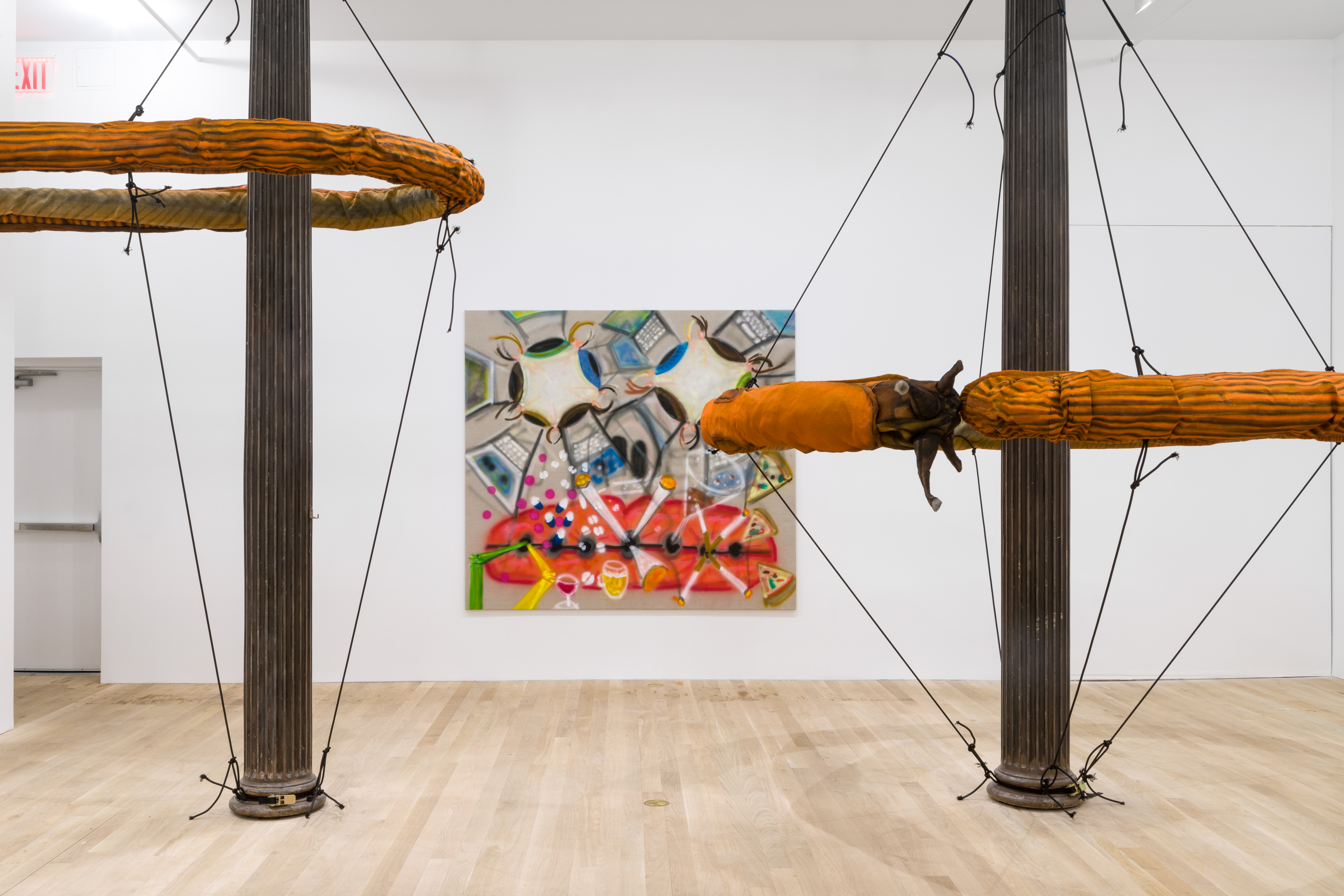 Two large, orange slug sculptures forming a circular ring shape around separate cast iron columns suspended by black elastic chords in a white walled gallery space with a square painting in the background.