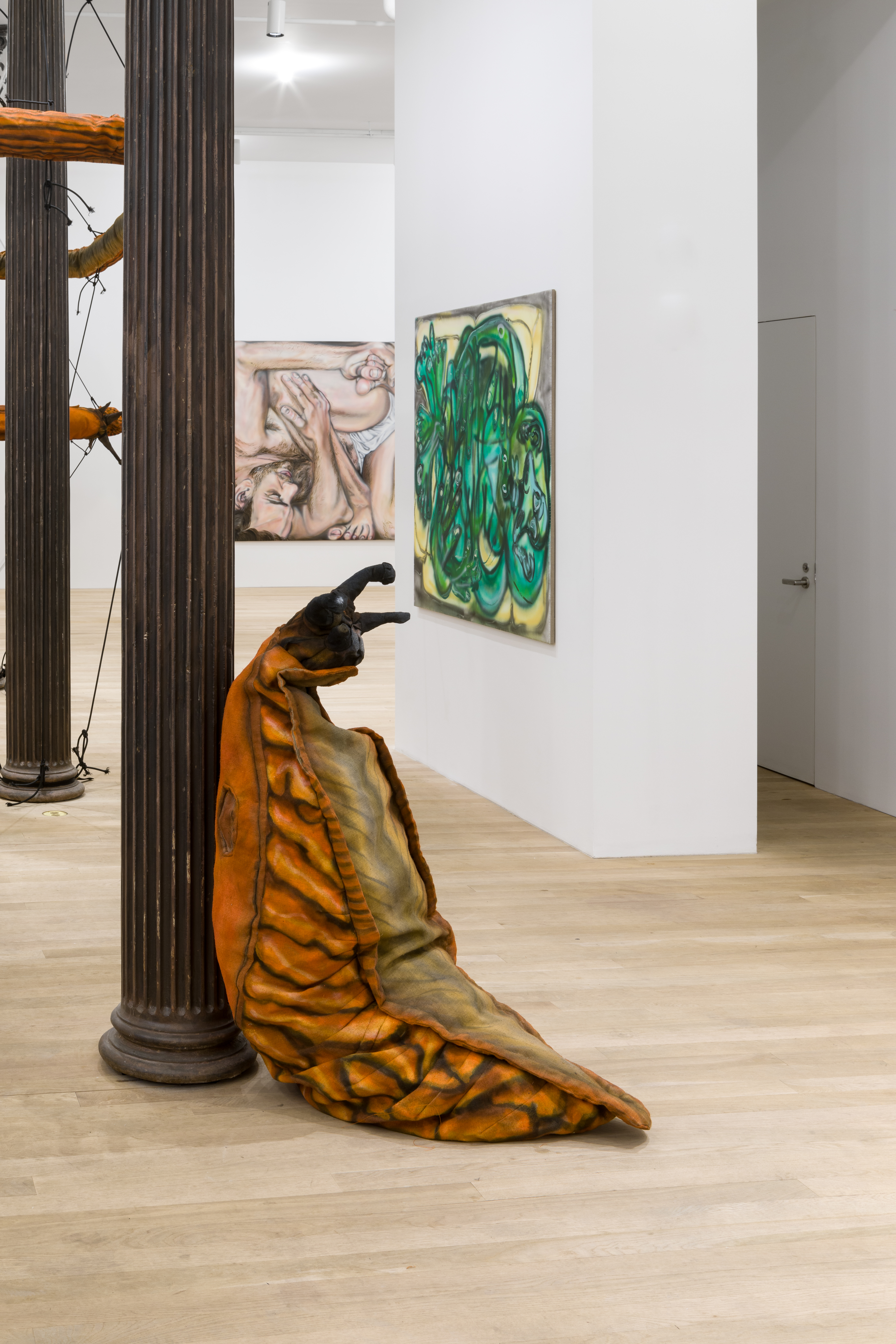 In the foreground is a slug sculpture leaning against a cast-iron column in a white-walled gallery space. In the background slug sculptures forming circular ring shapes can be partially visible around columns and two square paintings are visible on walls adjacent to one another.