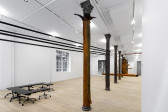 Large black straps wrap around some cast-iron columns, an orange, slug skins covers one of the cast-iron columns and other large, orange slug forms interact with the white-walled gallery space in the background. A black table sits in the corner and a figurative painting of an upright shark is in the back left corner.
