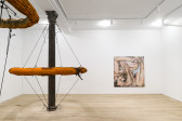A large, orange slug sculpture forming a circular ring shape around a cast-iron column suspended by eight elastic cords such that it encircles the column and rests at its vertical center. This is next to a square painting of a partially nude human figure in a white-walled gallery space. In the foreground the viewer can identify a partial view of a large, orange slug forming a circular ring near the ceiling.
