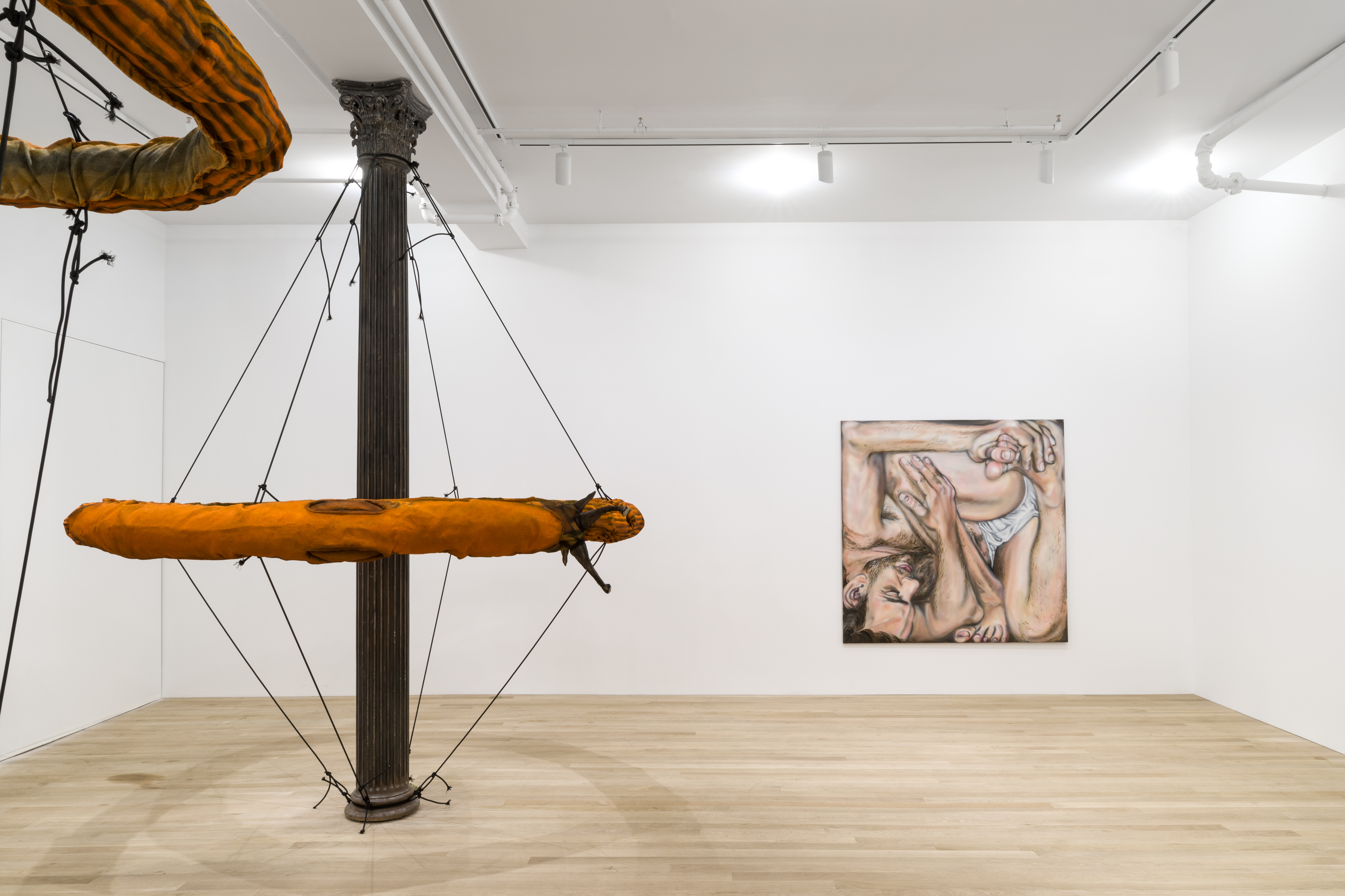 A large, orange slug sculpture forming a circular ring shape around a cast-iron column suspended by eight elastic cords such that it encircles the column and rests at its vertical center. This is next to a square painting of a partially nude human figure in a white-walled gallery space. In the foreground the viewer can identify a partial view of a large, orange slug forming a circular ring near the ceiling.