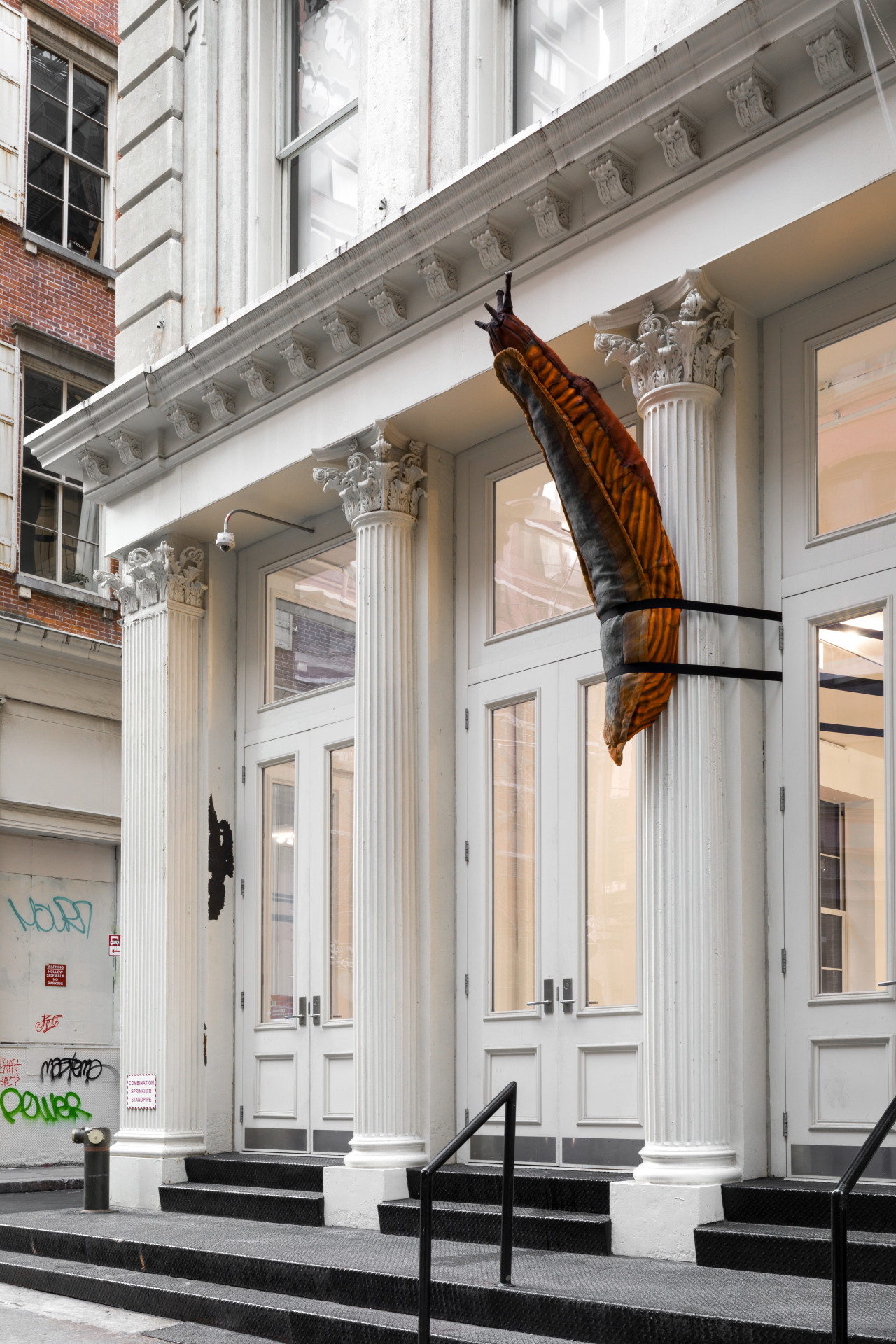 A large, orange and brown stuffed slug sculpture is strapped around a Greco-Roman style columns on the facade of a cast iron building. The slug’s antennae points upwards.