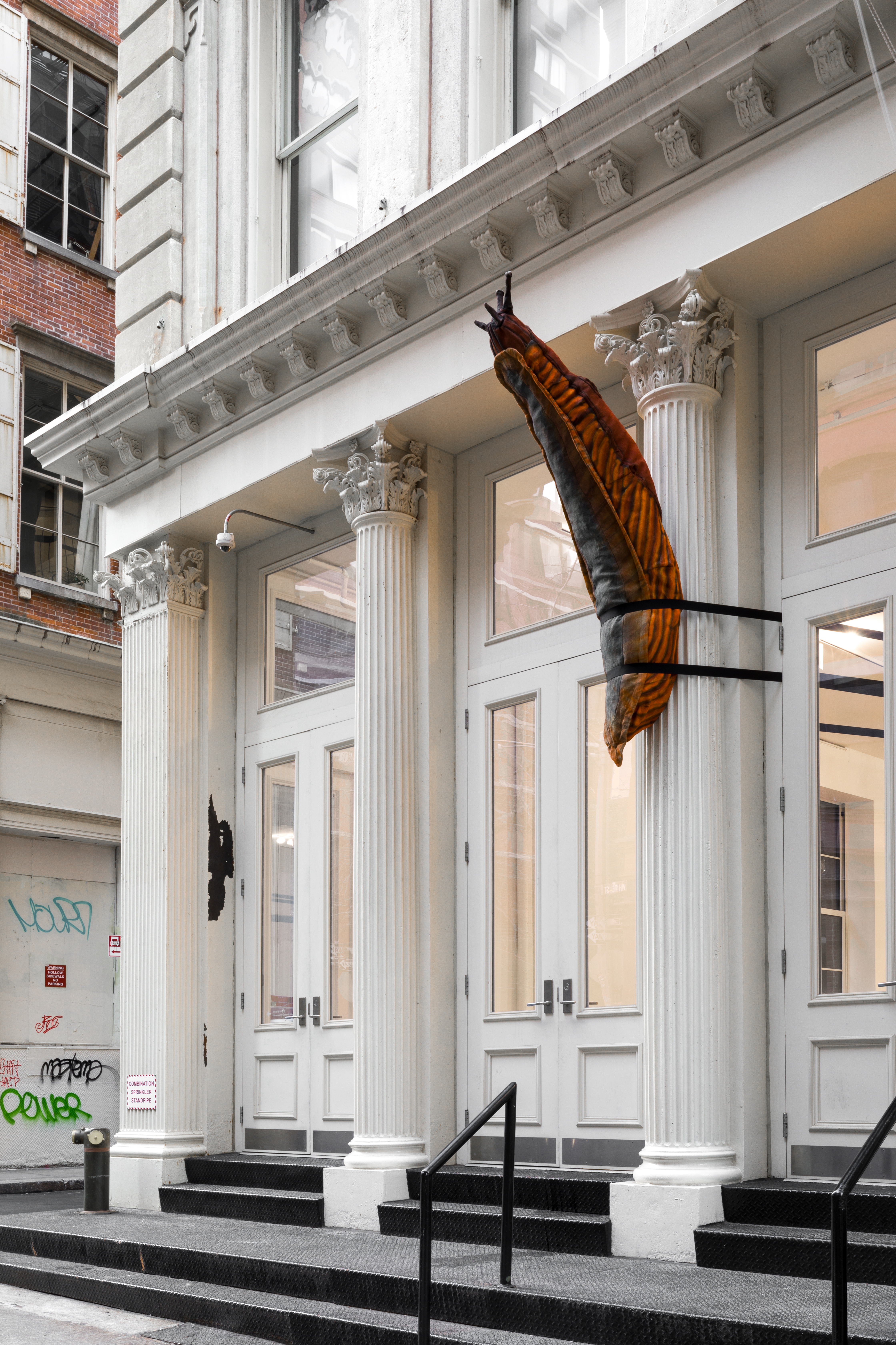 A large, orange and brown stuffed slug sculpture is strapped around a Greco-Roman style columns on the facade of a cast iron building. The slug’s antennae points upwards.