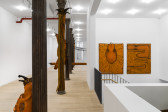 Large, orange slug forms are visible with the cast iron columns in a white-walled gallery with paintings of slugs hanging on the opposite side of the image behind a staircase.