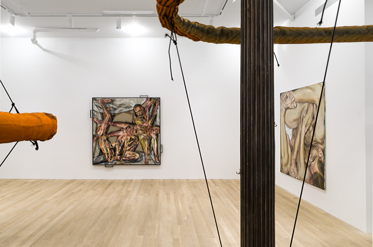 Partial view of two large orange slug sculptures forming a circular ring shape around cast-iron columns suspended by black elastic cords and two square paintings of the human form are on adjacent walls in a white-walled gallery space.