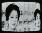 A distorted video still of a woman wearing pale makeup and lipstick. Her hair is carefully swept up and decorated with a hairpiece. A double of her image is positioned to her right. The background behind her displays several blurred kanji characters.