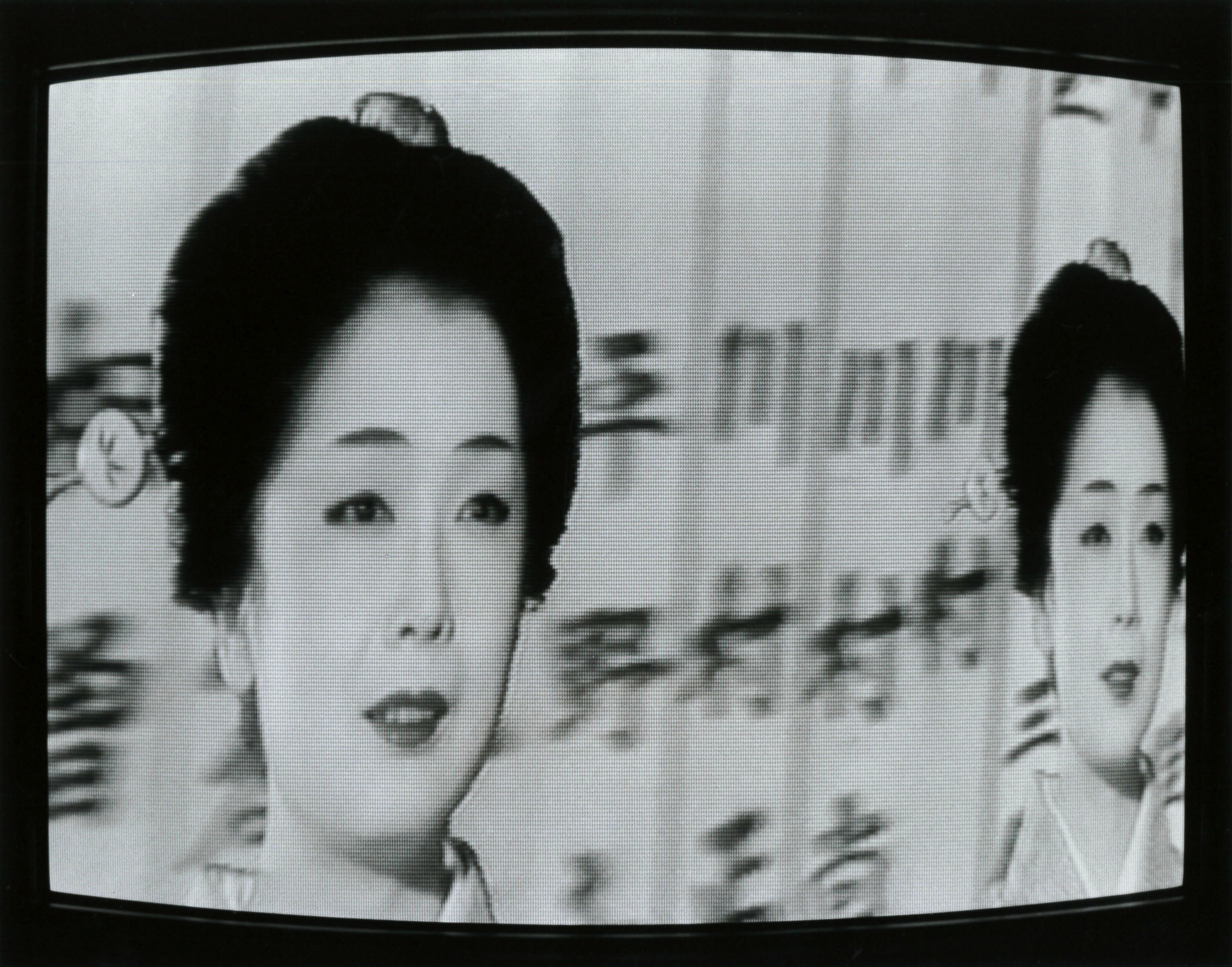 A distorted video still of a woman wearing pale makeup and lipstick. Her hair is carefully swept up and decorated with a hairpiece. A double of her image is positioned to her right. The background behind her displays several blurred kanji characters.