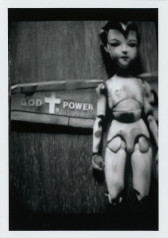 Black and white photograph of a doll lying on a wooden surface. To the left of the doll, white text on a metal surface reads, 