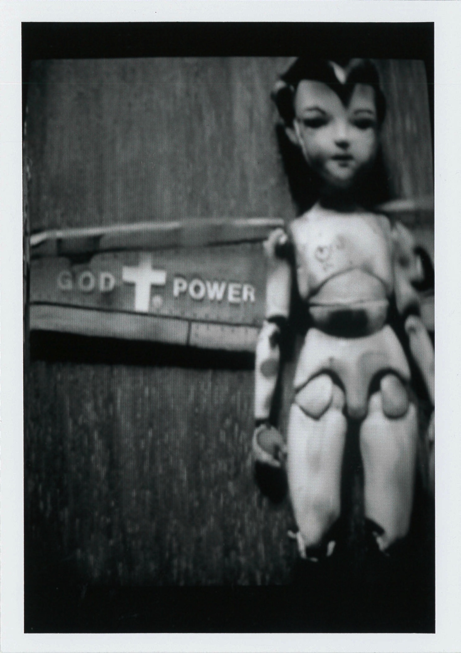 Black and white photograph of a doll lying on a wooden surface. To the left of the doll, white text on a metal surface reads, 