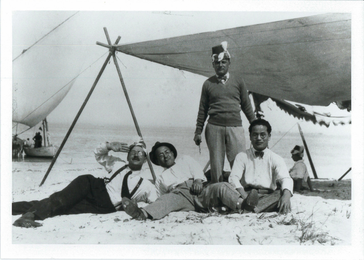 Black and white image of four men resting near a large, rigged tarp providing shade on a beach. Three of the men sit and recline on the sand, wearing white button-down shirts and trousers. The fourth man wears a hat and stands behind the men.