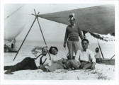 Black and white image of four men resting near a large, rigged tarp providing shade on a beach. Three of the men sit and recline on the sand, wearing white button-down shirts and trousers. The fourth man wears a hat and stands behind the men.