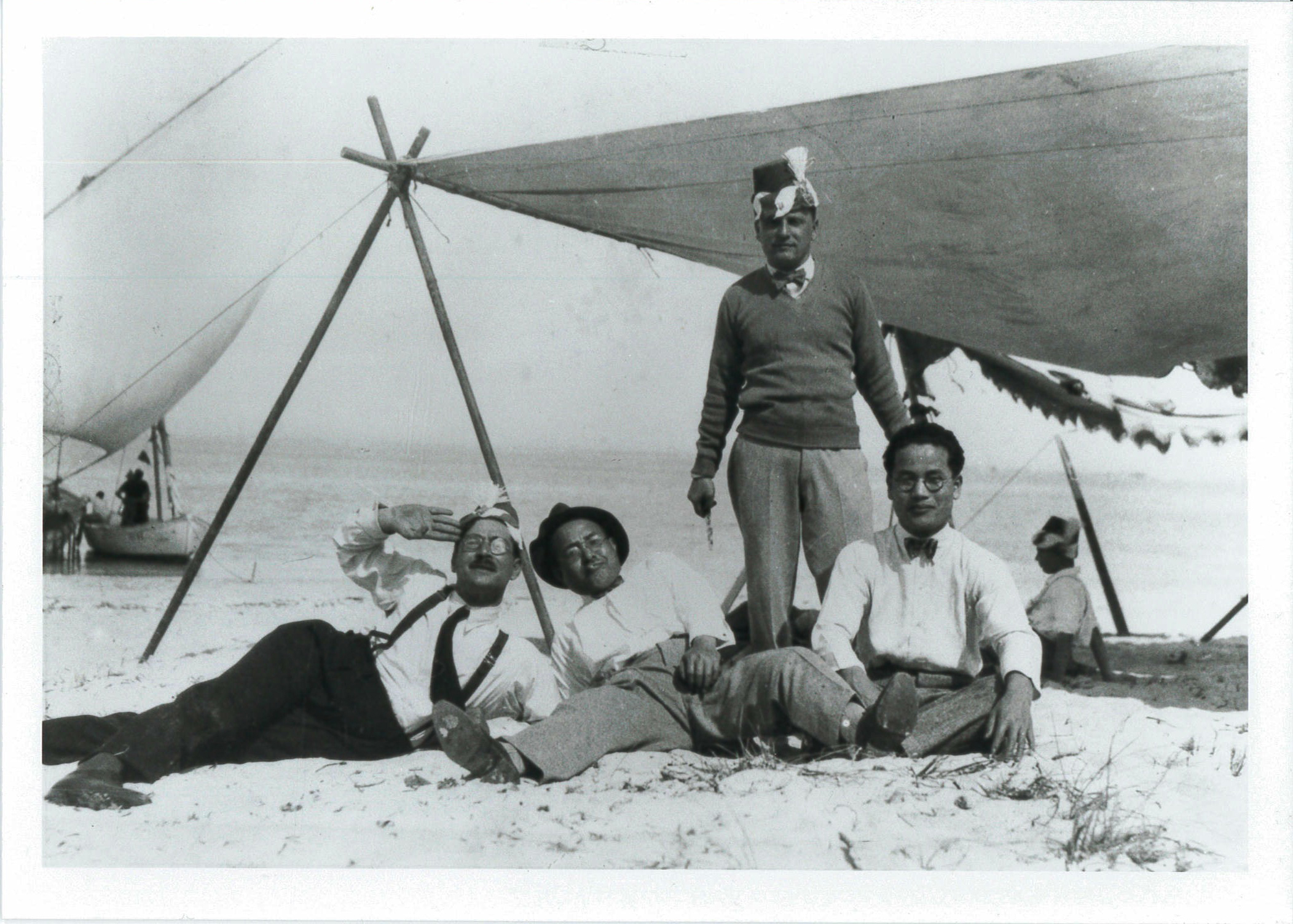 Black and white image of four men resting near a large, rigged tarp providing shade on a beach. Three of the men sit and recline on the sand, wearing white button-down shirts and trousers. The fourth man wears a hat and stands behind the men.