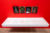 A color image of a close-up of a floating vitrine on a red wall, with four small paintings hung in a series above the vitrine. Inside the vitrine are six pages, with poems written on them and drawings of flowers decorating the pages. Thefour paintings above the poems depict Black Queer figures in darkened environments with gold, and colorful accents.