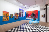 A color image of the corner of a gallery space. The walls are painted with blue accents, and there is a checkerboard floor. Large, vibrant paintings of Black Queer figures hang on the walls. Blue neon lights run along the frame of the window in the corner.