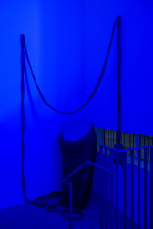 A color image of the stair landing inside a gallery. The whole image is illuminated in blue, and in the corner rests a large soft sculpture of hair clippers. A long cord connected to the clippers runs vertically up the wall and is draped to the other side. The outlet rests in the air, next to a windowsill.