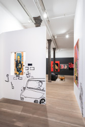A color image of a gallery space. To the left of the image, on the foreground, a white wall with a window and a drawing of a parked car. The driver is speaking with a figure that leans on the car. Through the window, a painting of three figures is visible. In the background, four works hang on a black wall. In the center of the room, a black couch and a table in the shape of a body.