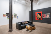 A color image of the corner of a gallery space. Two pillars run down the side of the space, inbetween them is a black leather couch with a soft sculpture of a figure laying down, wearing a durag, with a red PlayStation controller. In front of the couch is a table, comprised of a glass panel and a sculpture of a figure laying down with their backside raised. On the left wall is an arrangement of framed drawings. On the right wall is a large red painting, alongside more framed drawings.