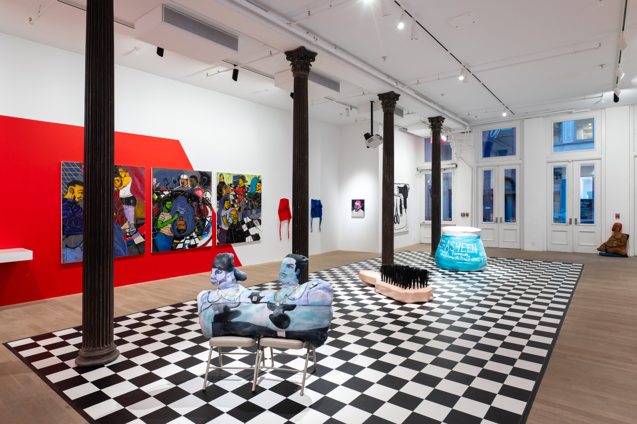 A color image of a large gallery room with columns running along the left side of the room. In the center is a checkerboard floor, with three large sculptures of two figures, a hairbrush, and a bottle of hair gel placed on it. The left wall is painted red, and large paintings and durags are hung across it.