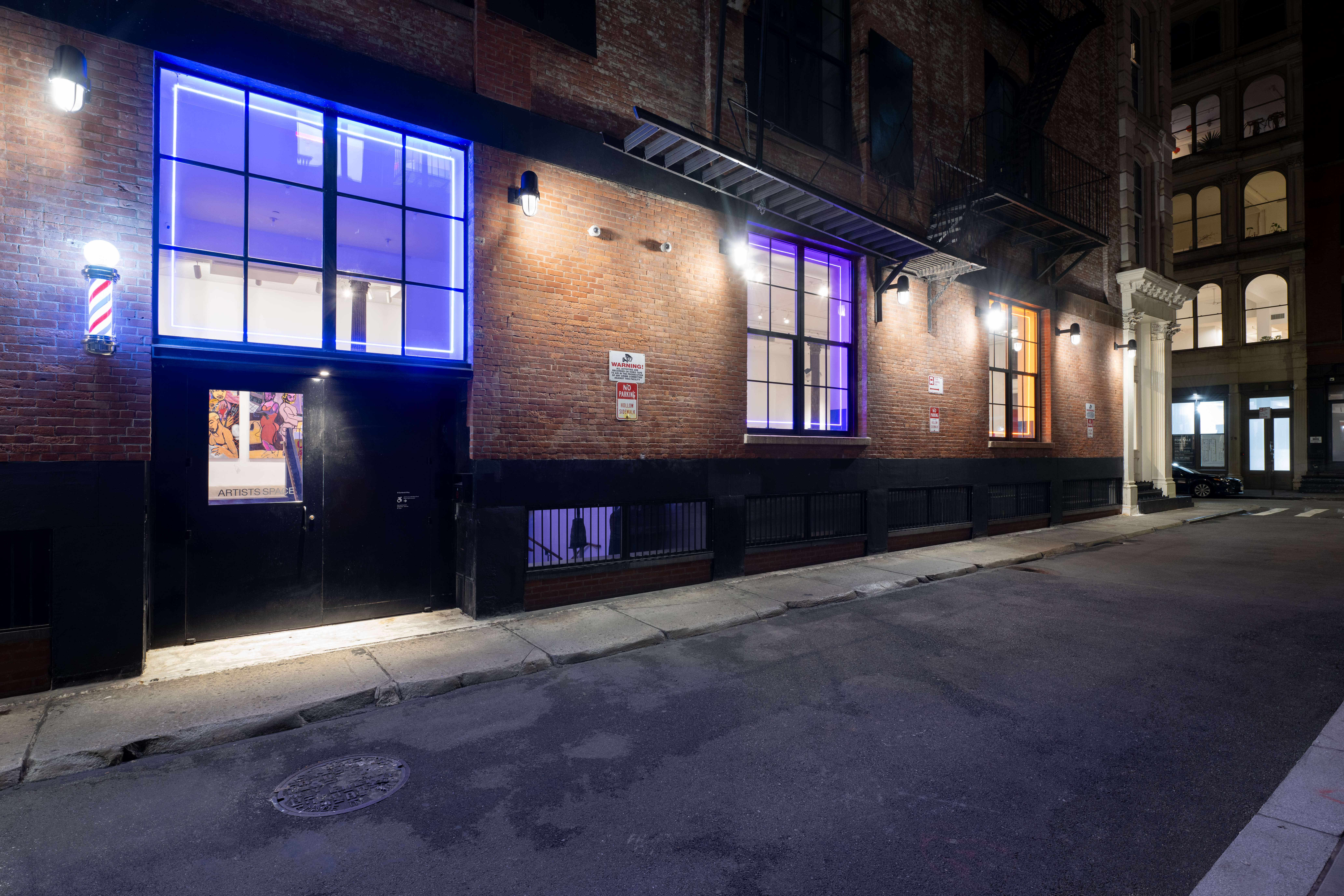 A color image of the outside of a brick building. Three large windows running along the wall are each illuminated with neon lights. On the left side is a door to a gallery space, through which can be seen some large, colorful paintings hanging inside.