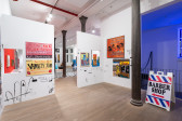 A color image of a gallery space divided by two walls, with columns running down the middle. There are multple paintings and drawings hanging on the walls. In the center of the back room, a black-and-white checkered floor. To the right, an A-frame sign with the words 