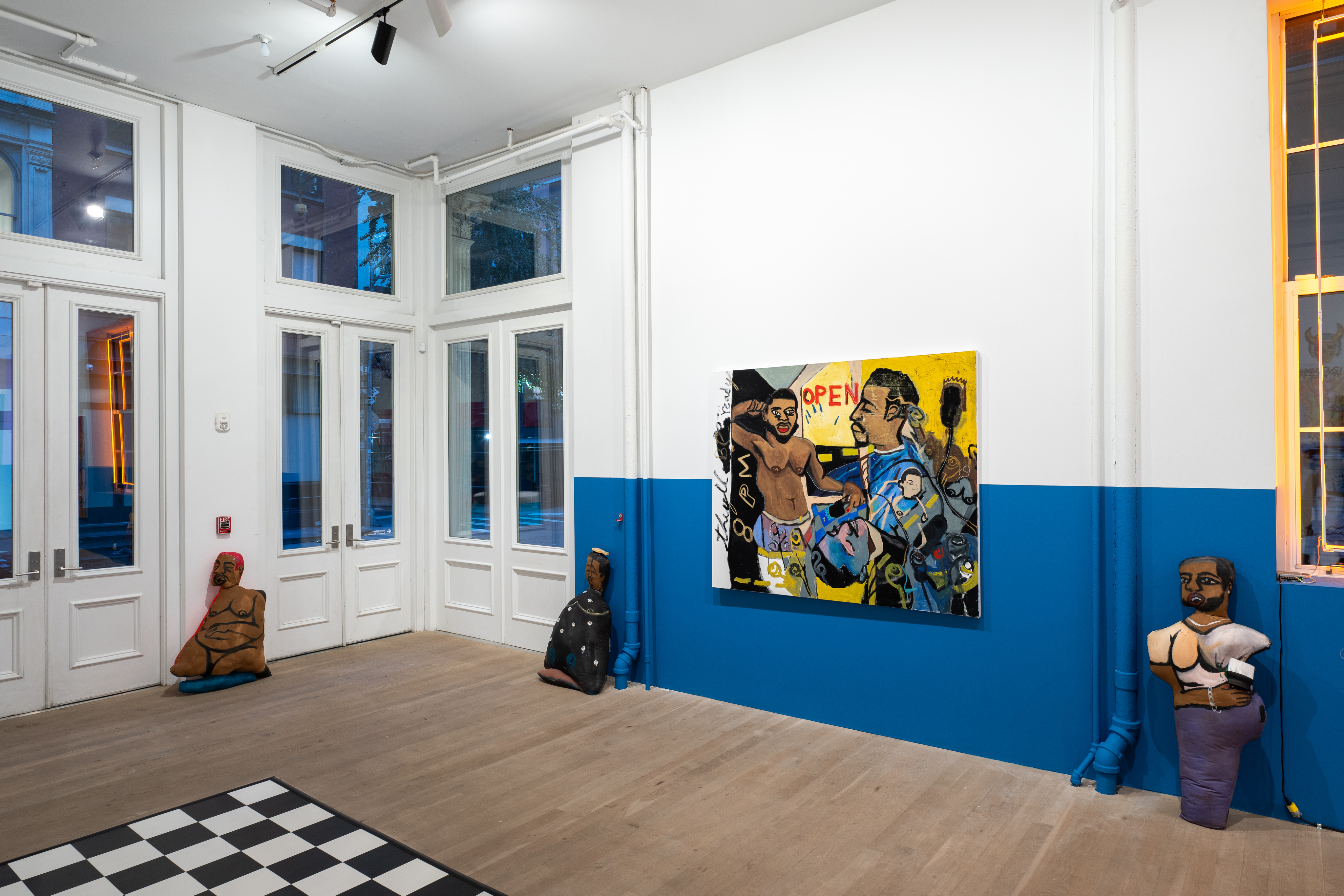 A color image of the corner of a gallery space. Half of the right wall has been painted blue, and a yellow painting of Black Queer figures in a barbershop is hung on the wall. Three soft sculptures are placed, evenly spaced apart along the floor, all depicting different figures.