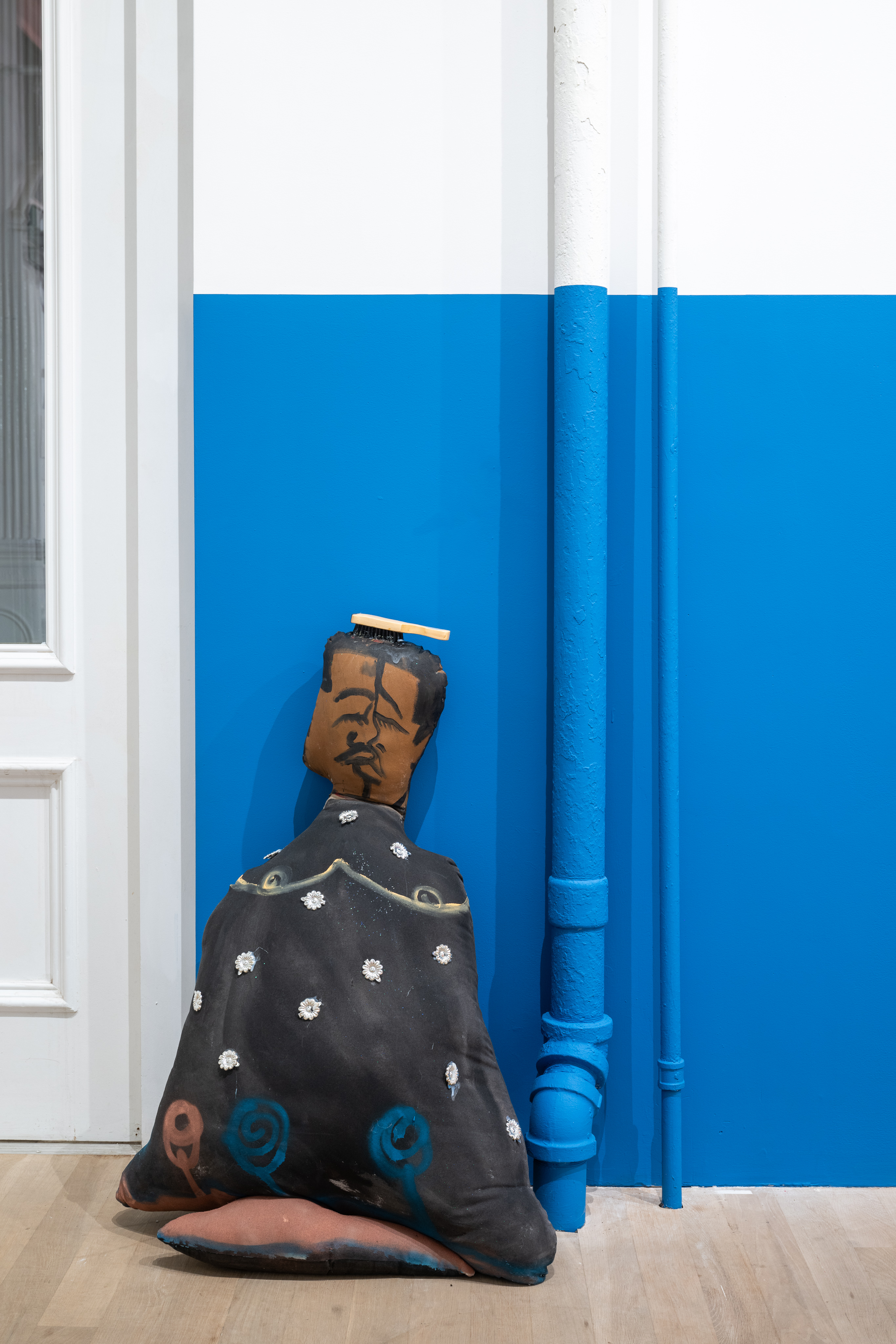 A color image of a soft sculpture, placed on the floor and leanning against a blue and white wall. The sculpture depicts a Black Queer figure dressed in black, with accents of white, blue and yellow. On top of their head is a wooden brush.