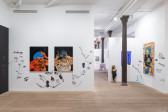 A color image of a gallery space, with large paintings of Black Queer figures placed on white walls. Drawings in black accent the walls and complement the paintings. Past the left wall is a corridor leading into the next room. In the distance is a white sign on the floor, reading 