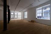 Installation view of a gallery space with sunlit windows. On the right, a row of colums extends into the background. A wooden bench is pushed against the wall on the left under a windowsill with several books.