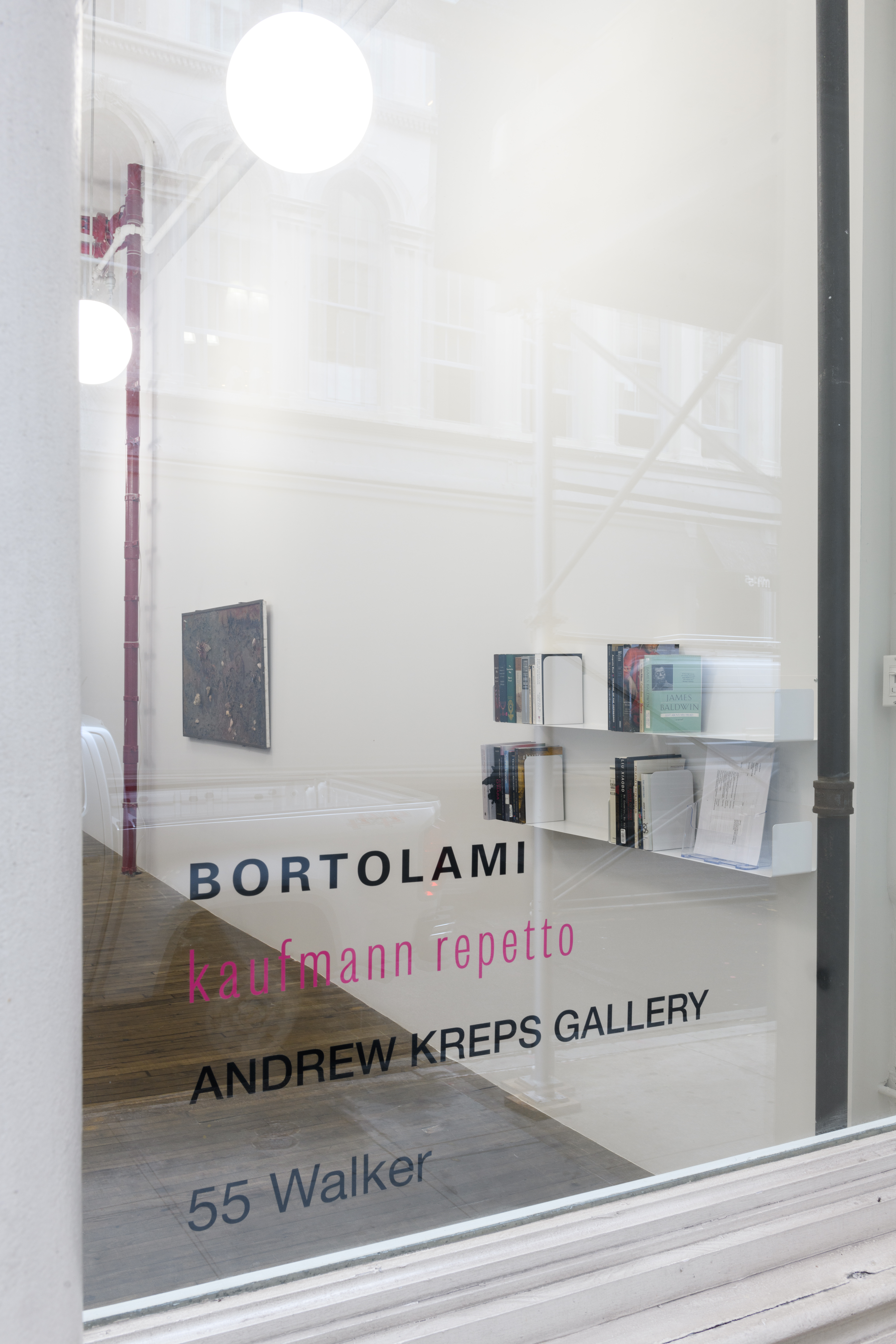 Looking into a gallery space through a window, on the right wall two shelves are filled with books. Further in the background a dark rectangle hangs on the wall. On the windowpane the words 