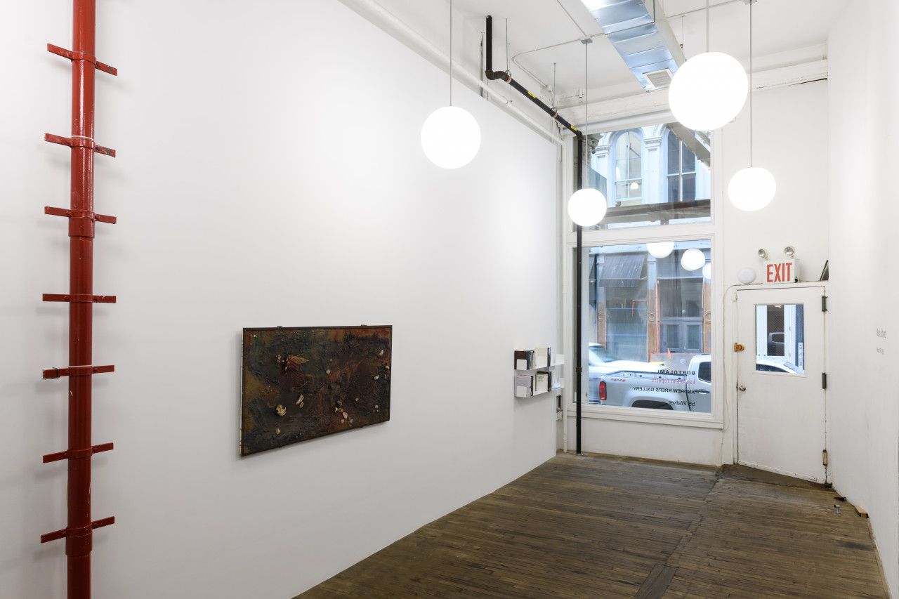 Installation image of a gallery space with a window looking out onto a street. On the left, a red pole with ladder rungs, a rectangular collaged artwork, and two shelves holding books and papers are visible.