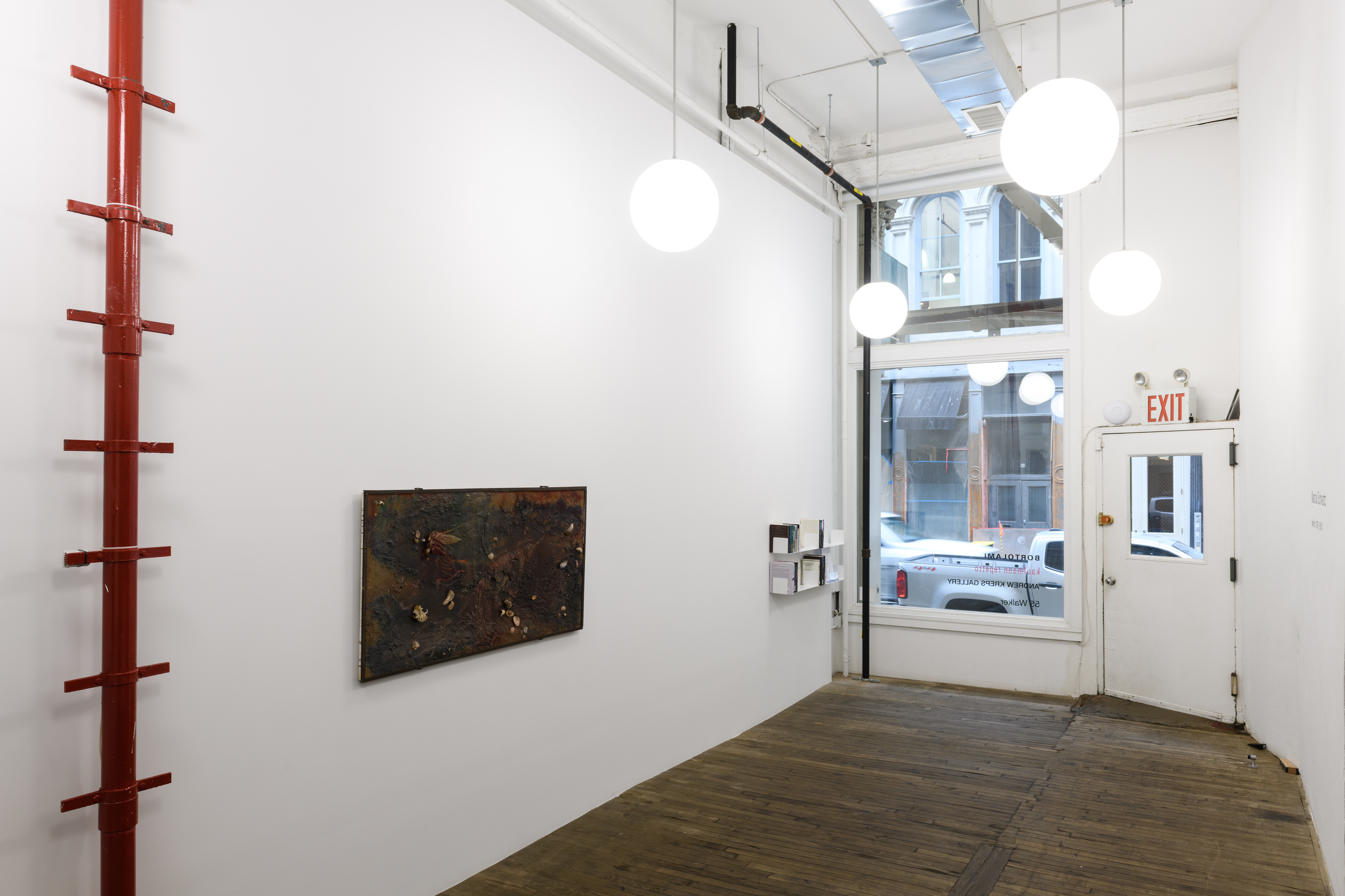 Installation image of a gallery space with a window looking out onto a street. On the left, a red pole with ladder rungs, a rectangular collaged artwork, and two shelves holding books and papers are visible.
