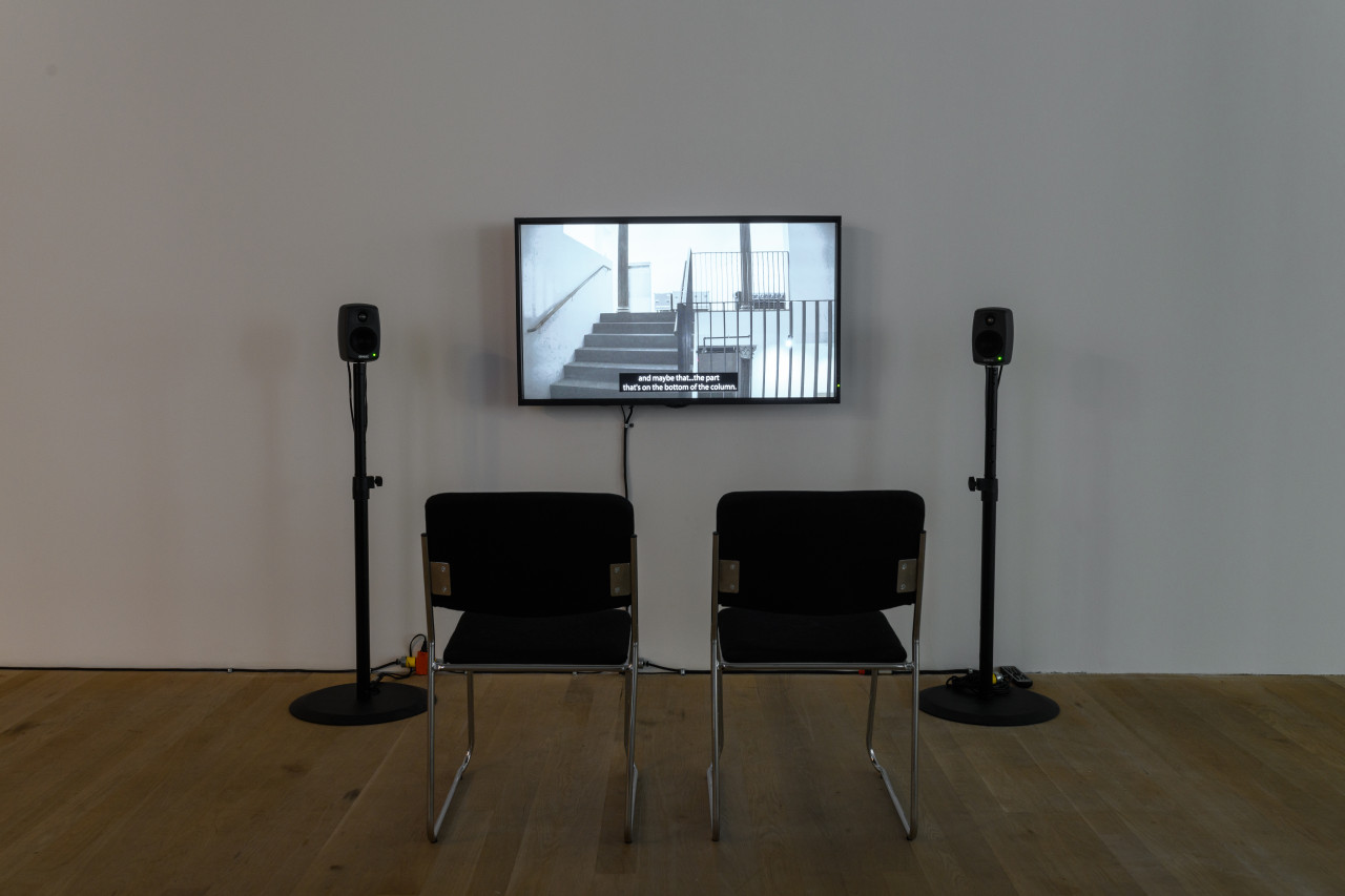 Installation view of a video work. A television monitor is attached to the wall, flanked by two speakers on narrow stands. In front of the monitor are two black chairs with silver metal legs.